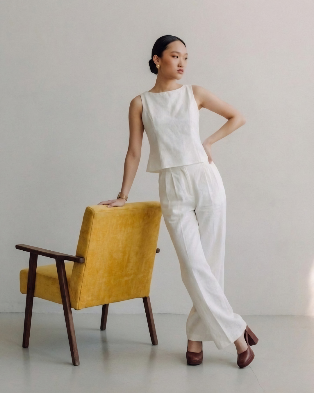 Woman in a white outfit standing next to a yellow chair against a plain background